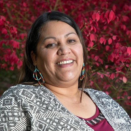 A smiling woman wearing colorful earrings poses in front of vibrant pink flowers.