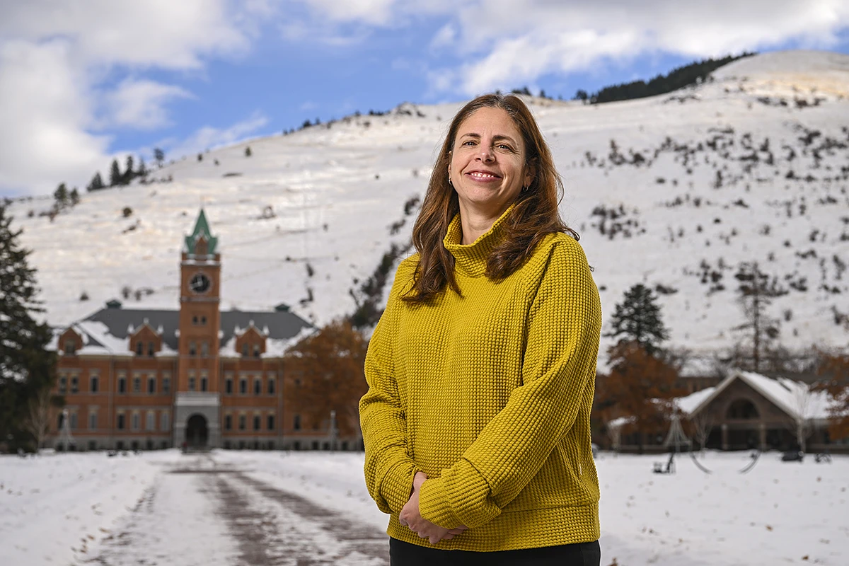 A smiling woman in a yellow sweater stands outdoors in front of a snow-covered landscape with a large building in the background.