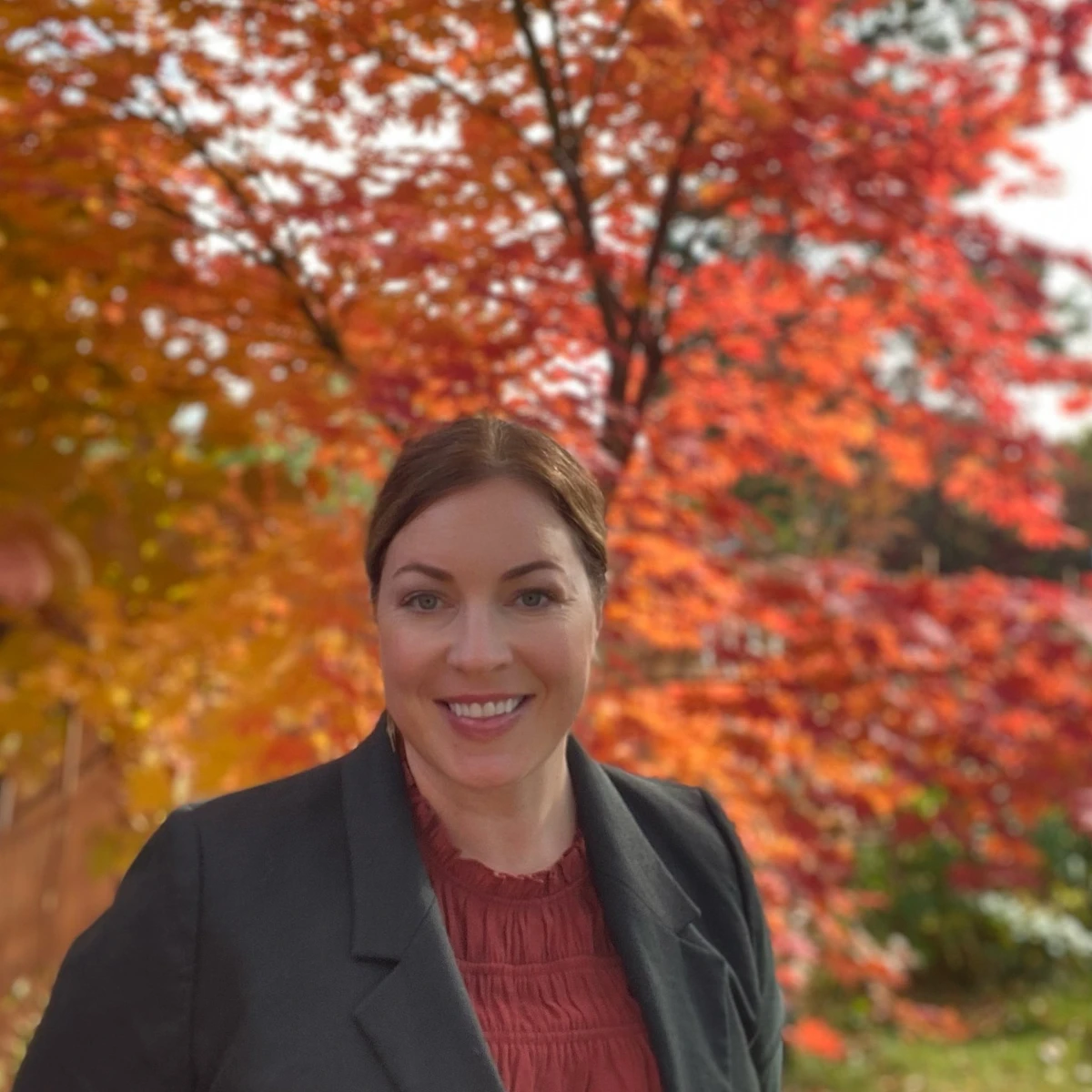 A woman with her hair pulled back stands smiling in front of a vibrant autumn tree with red and orange leaves.