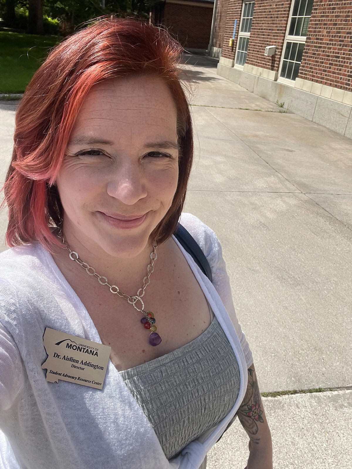 A woman with short red hair and a name tag, wearing a grey shirt and a white cardigan, smiles outdoors near a brick building.