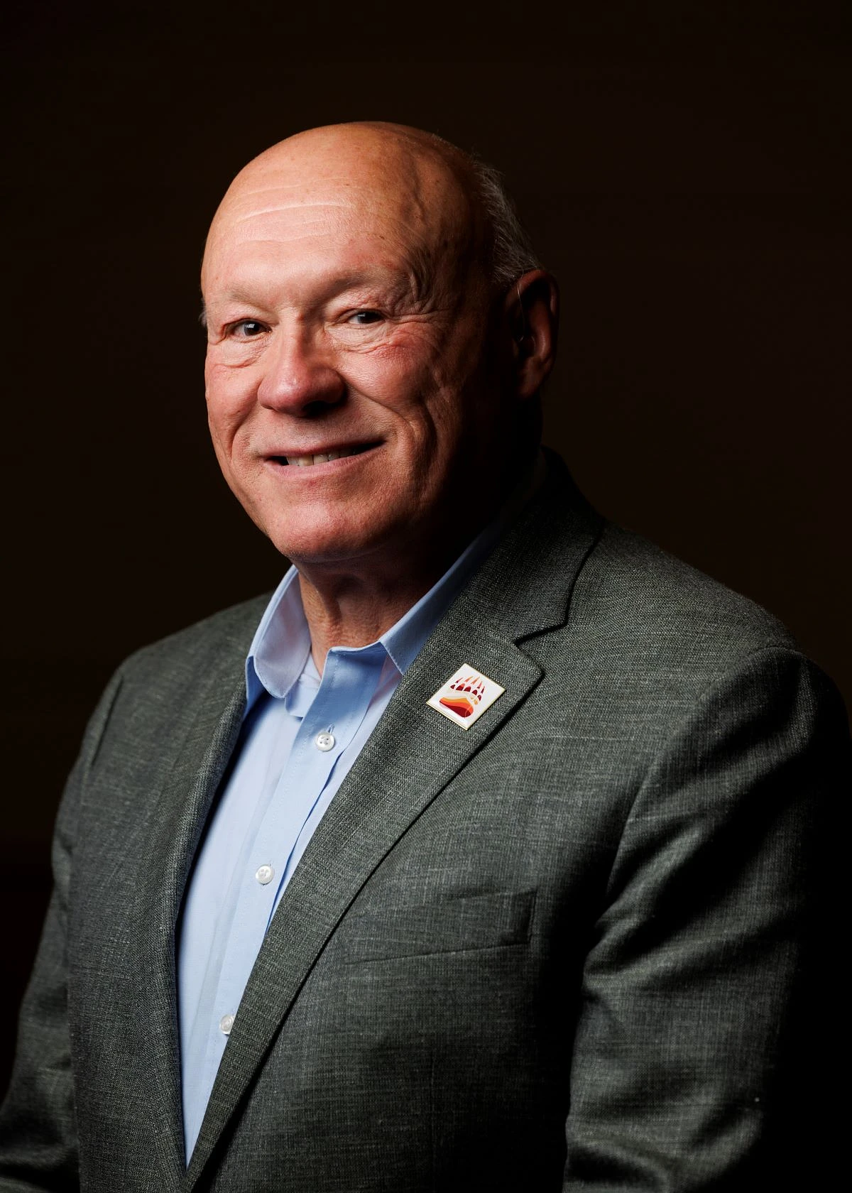 A smiling bald man in a suit and tie with a pin on his lapel poses against a dark background.