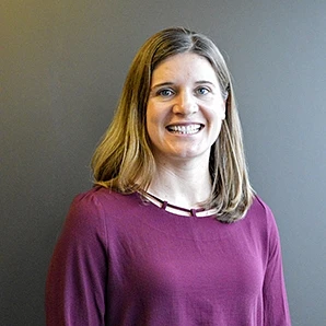 A woman with shoulder-length hair is smiling in front of a plain background, wearing a maroon top.